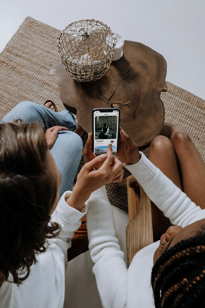 Two women sitting by a wooden table, using a smartphone app for online dating advice and profiles.