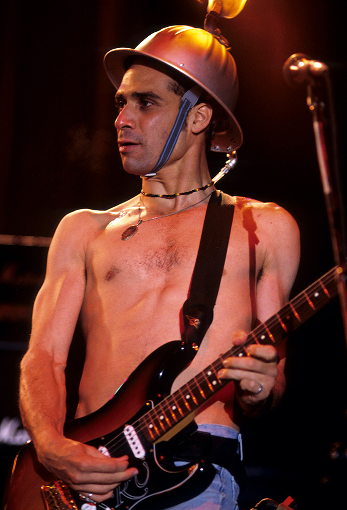 Shirtless former Red Hot Chili Peppers guitarist wearing helmet, playing electric guitar on stage under warm lighting.