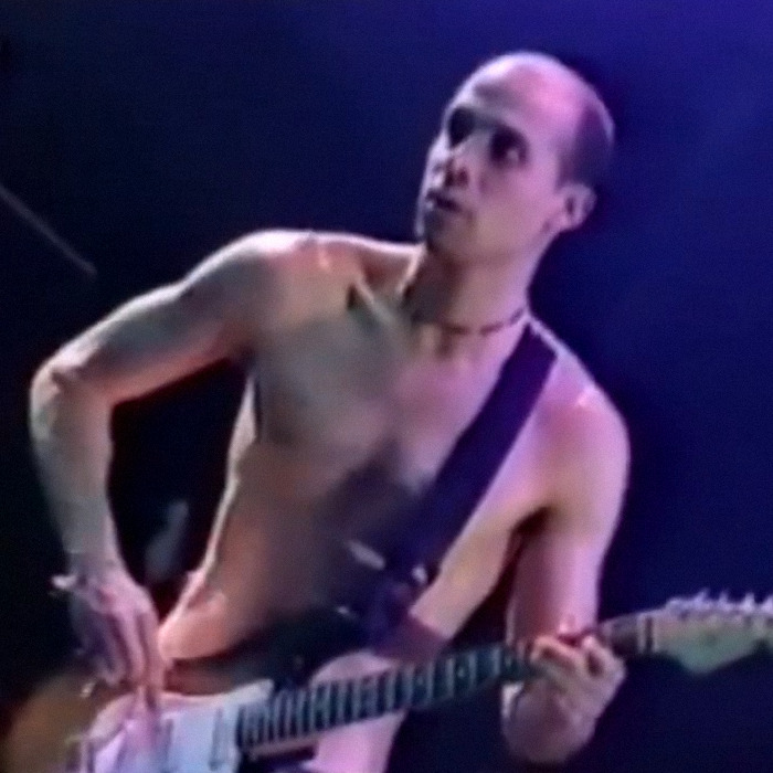 Former Red Hot Chili Peppers star playing electric guitar shirtless on stage under purple lighting at a live performance