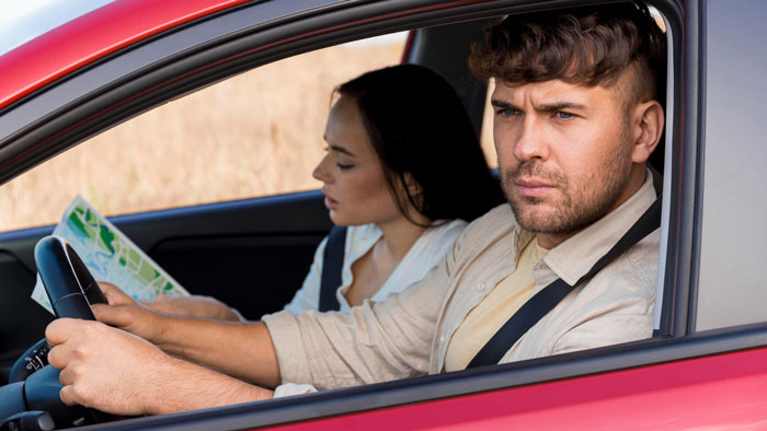 Man looking frustrated while driving car with woman reading map beside him, reflecting husband sick of hosting relatives.