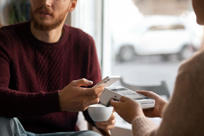 Man in a maroon sweater making a contactless payment with a smartphone while holding a coffee cup indoors.