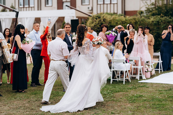 Bride walking in elaborate wedding dress outdoors with guests capturing the moment on phones during wedding ceremony.