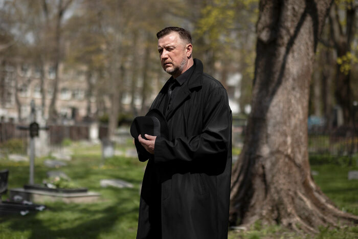 Man in black coat holding hat solemnly at a funeral, reflecting on mother, partner, and ex-husband relationships.