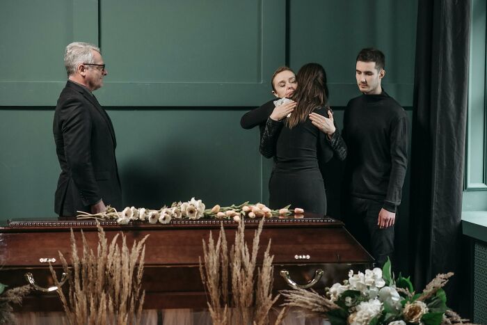 Mourners including mother, partner, and ex-husband comforting each other during a funeral beside a closed casket.
