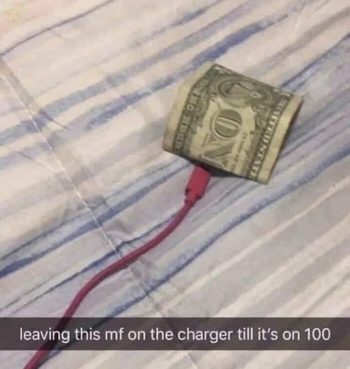 Rolled dollar bill attached to a phone charger on a bed, humorous meme about charging money to 100 percent.