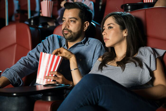 Couple sitting in a movie theater, focused on screen, woman holding popcorn, illustrating unhinged stalking stories theme.