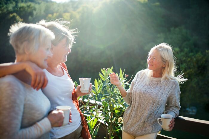 Three older women talking and holding mugs outdoors, sharing stories about near-death experiences in bright sunlight.