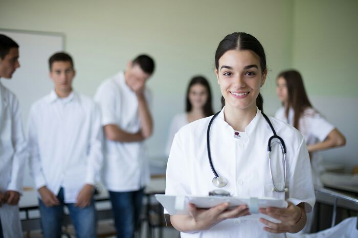 Young female doctor smiling with clipboard and stethoscope in clinical setting with medical team in background, health hacks concept