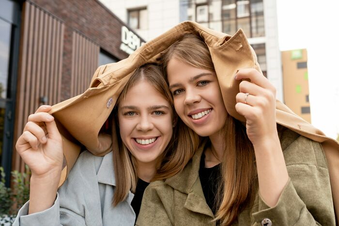 Two young women smiling under a coat outdoors.