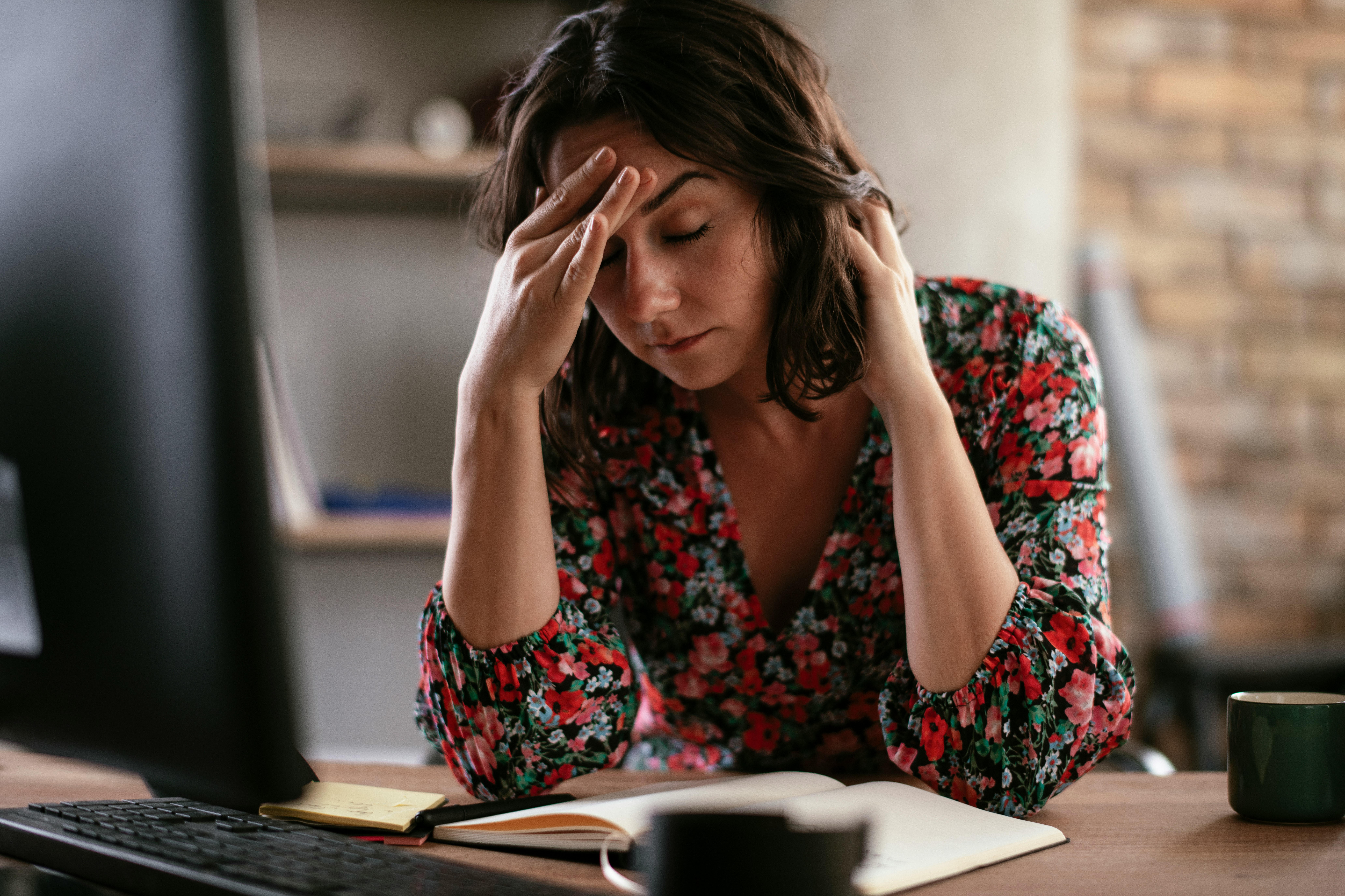 Tired woman in floral blouse rubbing forehead at desk, illustrating workplace perfume ban and employee allergies conflict.