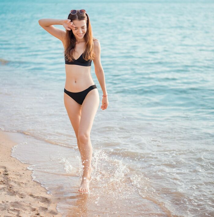 Young woman in black bikini walking on the beach with water splashing, unrelated to wedding guests or bride and groom regrets.