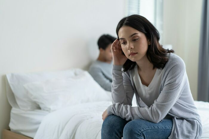 A woman looking sad and distant, sitting on the edge of the bed while a man sits blurred in the background, depicting cheating signs in women.