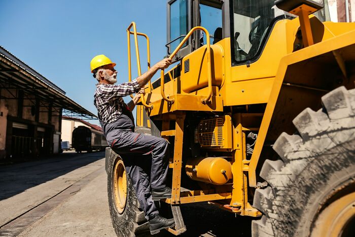 An older construction worker wearing a hard hat climbs a large yellow heavy machinery vehicle at a worksite.