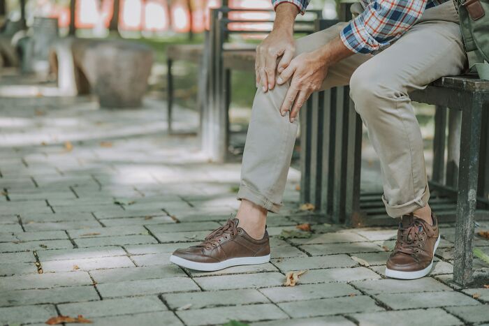 Person sitting on a bench outdoors, holding their knee in discomfort, illustrating effects of medical neglect today.