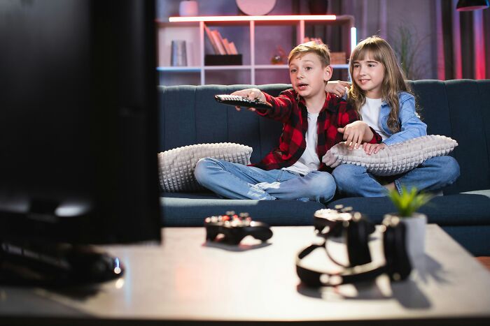 Two children having a playdate watching TV together on a couch, surrounded by gaming controllers and virtual reality headsets.