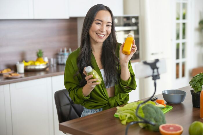 Young woman smiling in kitchen holding healthy green and orange juices with fresh vegetables on table promoting healthy habits.