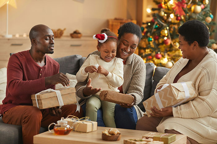 Family exchanging gifts by a decorated Christmas tree, capturing a joyful moment with kid&rsquo;s photos and a dog picture prank.