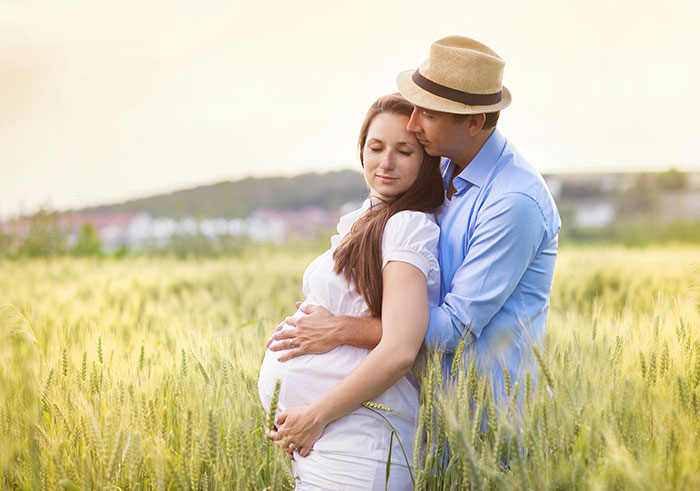 Pregnant wife embraces husband in wheat field while discussing baby name inspired by childhood dog Lassie.
