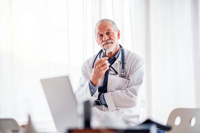 Elderly male doctor with stethoscope and glasses reflecting on near-death experiences in a bright medical office.