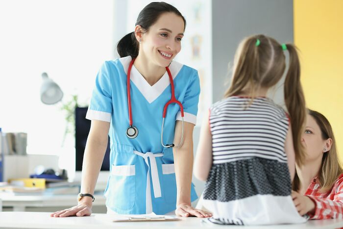 A nurse in blue scrubs with a red stethoscope talking to a young girl, illustrating strange health hacks shared by patients.