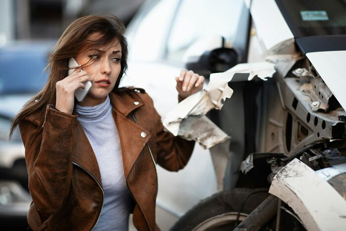 Woman in brown jacket making a phone call beside a damaged white car, showing moments the universe protected people from harm.