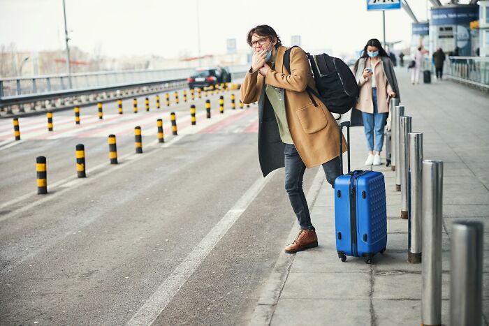 Young traveler with blue suitcase wearing a brown coat and face mask, standing on the sidewalk near the road.