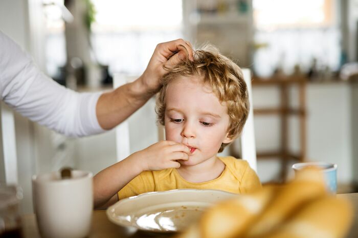 Young child eating at a table with an adult hand gently touching their hair, reflecting childhood rules and early development moments.