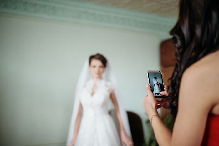 Bride posing in wedding dress while guest takes a photo on smartphone, illustrating wedding guests causing regret moments.