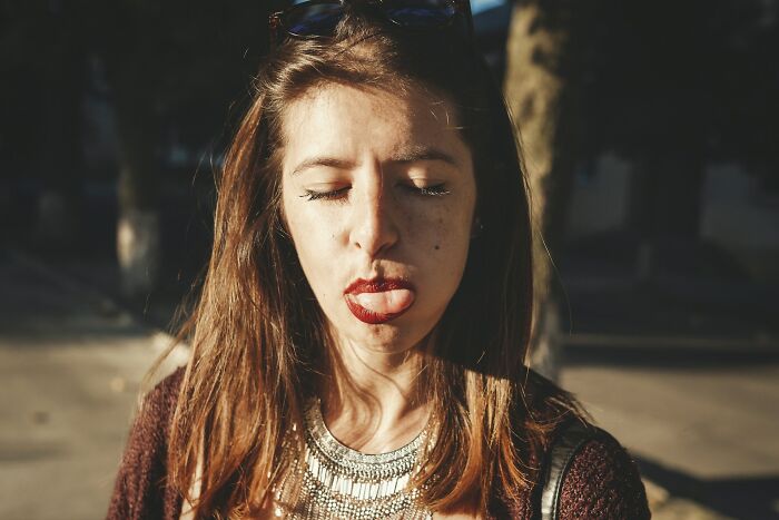 Young woman with eyes closed and tongue out, outdoors in sunlight, expressing disbelief and frustration about common propaganda truths.