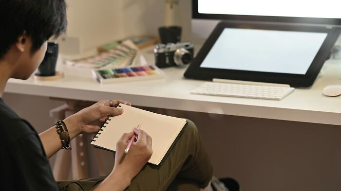 Person sitting near a desk with a blank notebook and pen, reflecting on job loss due to AI impact.