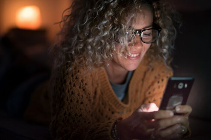 Woman with curly hair and glasses lying on couch at night, smiling while checking phone showing possible cheating signs in women.