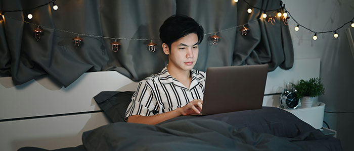Young man in striped shirt in bed at night, looking concerned while using laptop, hinting at creepy secret photos discovery.