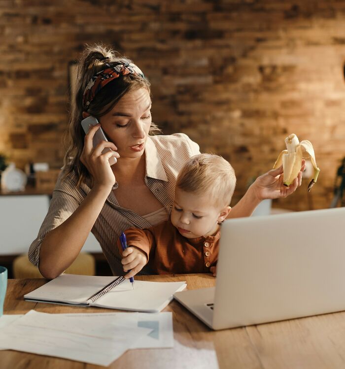 Female professor multitasking at home with child, eating banana, working on laptop, and talking on phone discussing evidence-based decision.