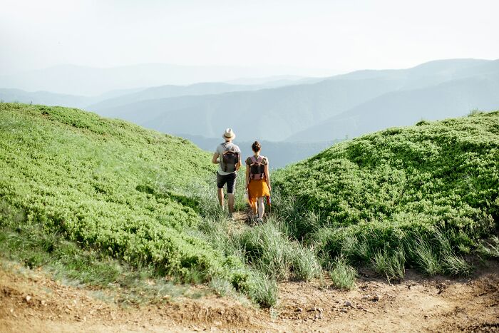 Two hikers with backpacks walking on a mountain trail surrounded by green hills and distant blue mountains.