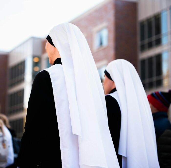 Two nuns wearing white veils and black habits standing outdoors in an urban setting at a funeral event.