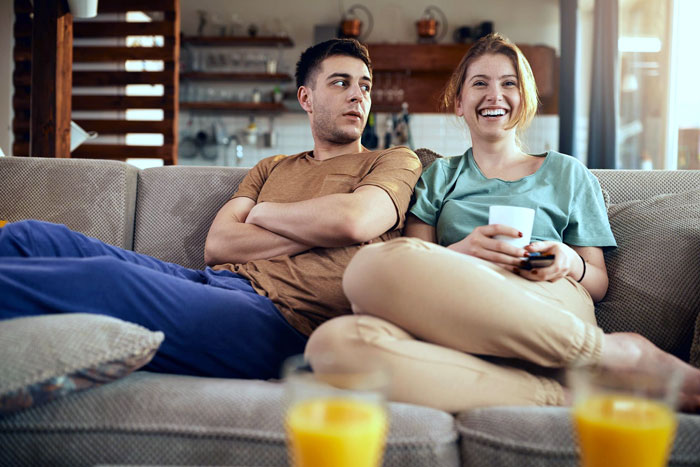 Couple sitting on couch with boyfriend looking concerned and girlfriend smiling, tense moment about trip with female friend.
