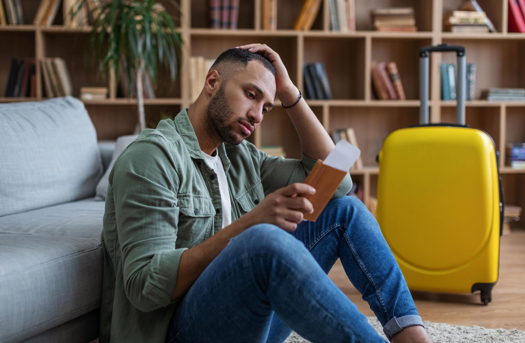 Man reconsidering relationship while holding travel document near yellow suitcase in living room with bookshelves.