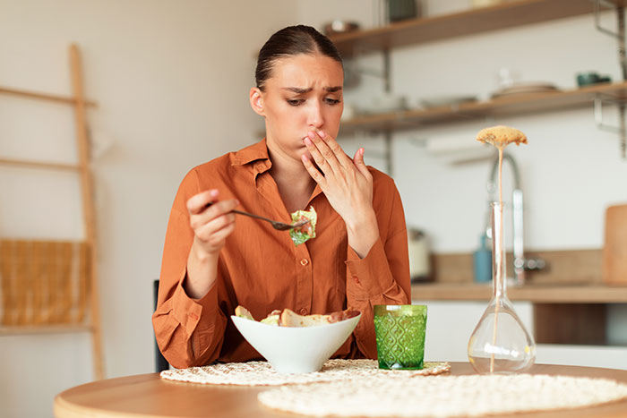 Woman looking sick and hesitant while eating homemade food at a kitchen table, concerned about getting sick repeatedly. Woman looking sick and hesitant while eating homemade food at a kitchen table, concerned about getting sick repeatedly.