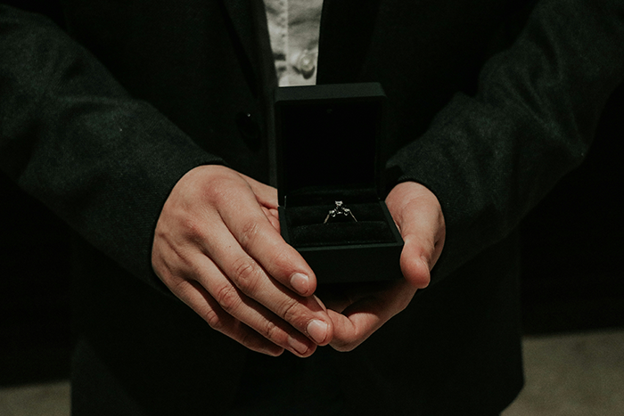 Man in dark suit holding an engagement ring box, symbolizing a proposal with an open marriage twist.