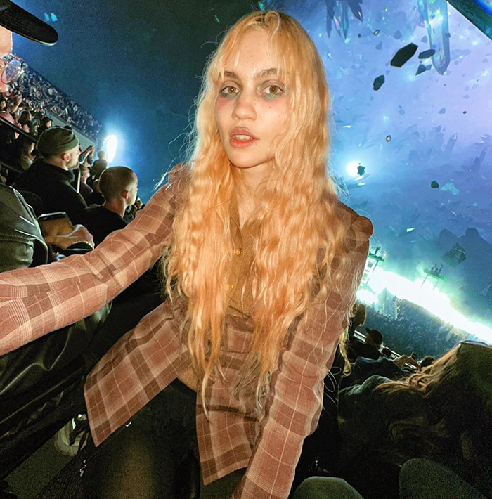 Grimes with long blonde hair wearing a plaid jacket at an event, showing off her ringworm face tattoo.