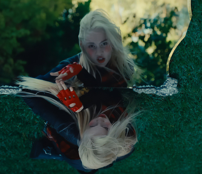 Woman with platinum blonde hair wearing red gloves, bending over a reflective surface, showcasing a ringworm-inspired face tattoo.