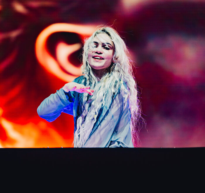 Grimes performing on stage with long blonde hair and colorful background, showcasing her face tattoo and ringworm style.