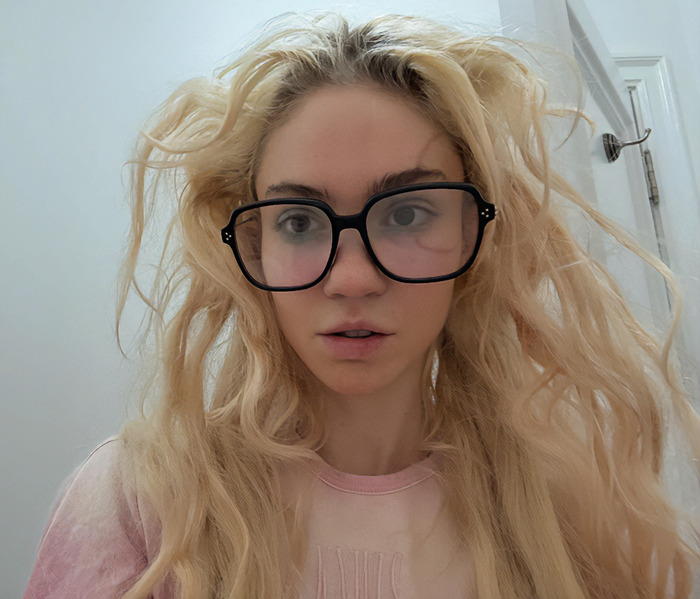 Close-up of a woman with messy blonde hair and large glasses, showcasing a unique ringworm face tattoo.