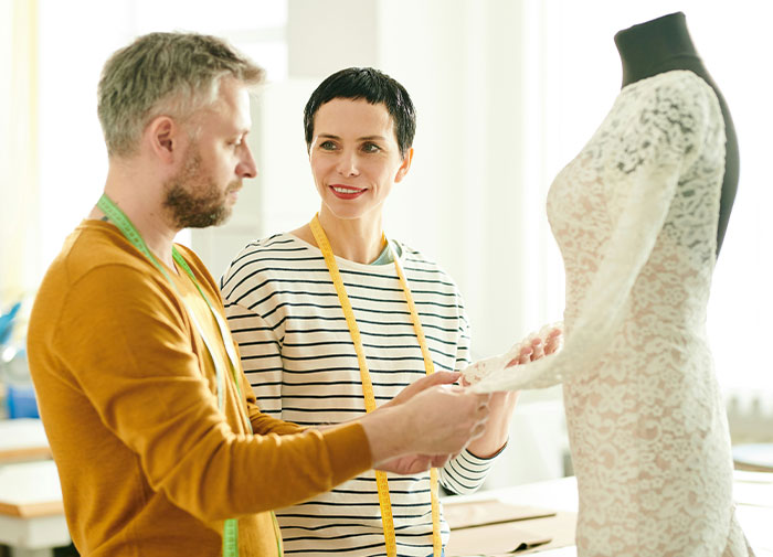 Woman and man with measuring tapes examining lace fabric on a wedding dress mannequin for guest wearing own wedding dress guidance
