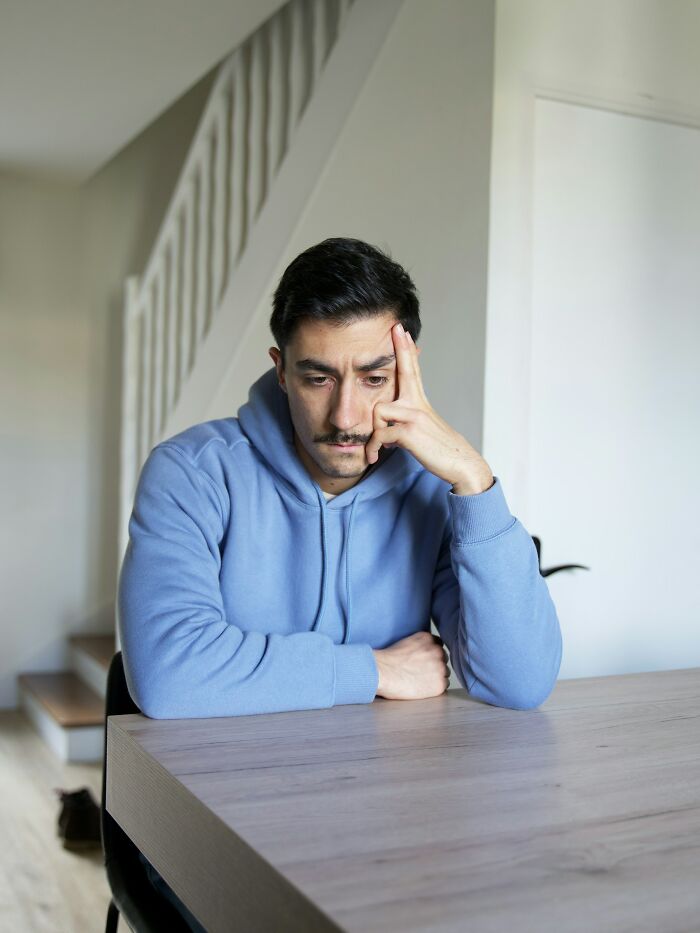 A man in a blue hoodie sitting alone at a table, looking thoughtful and sad after heartbreaking betrayal by close friends.