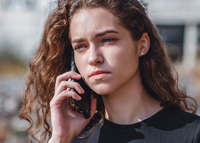 Young woman with curly hair looking uncomfortable while talking on phone outdoors, reflecting fragile masculinity issues.
