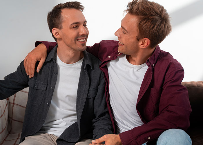 Two men sitting closely on a couch, smiling and showing a moment of connection related to masculinity so fragile concept.
