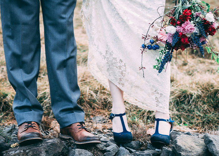 Close-up of bride and groom&rsquo;s feet on rocky ground, bride holding colorful bouquet at a Halloween wedding event.