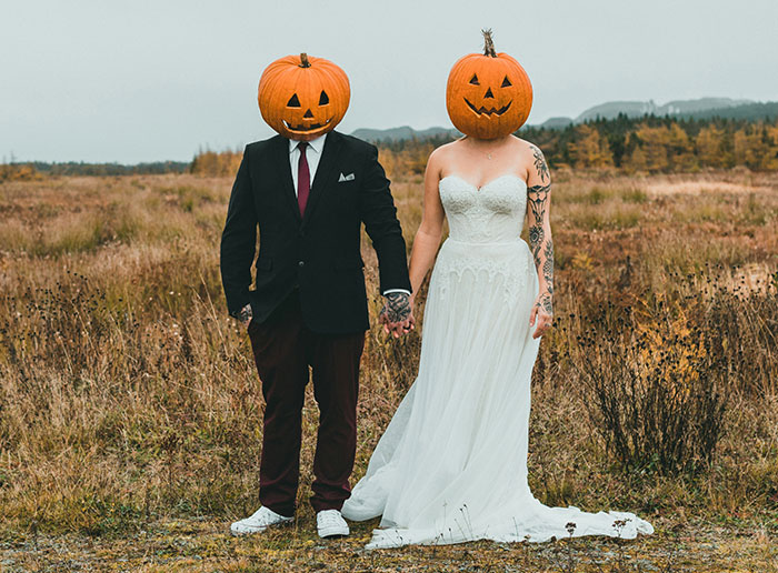 Bride and groom wearing carved pumpkin heads at a Halloween wedding in an open field with autumn colors.