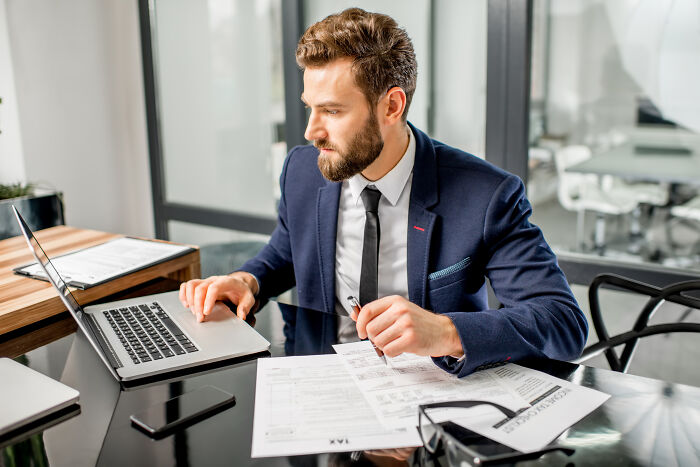 Young accountant in a navy suit working on tax documents and laptop at a modern office, showing accountants' engaging stories.